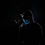 Silhouette of a person in a hoodie wearing a glowing LED mask against a dark background.